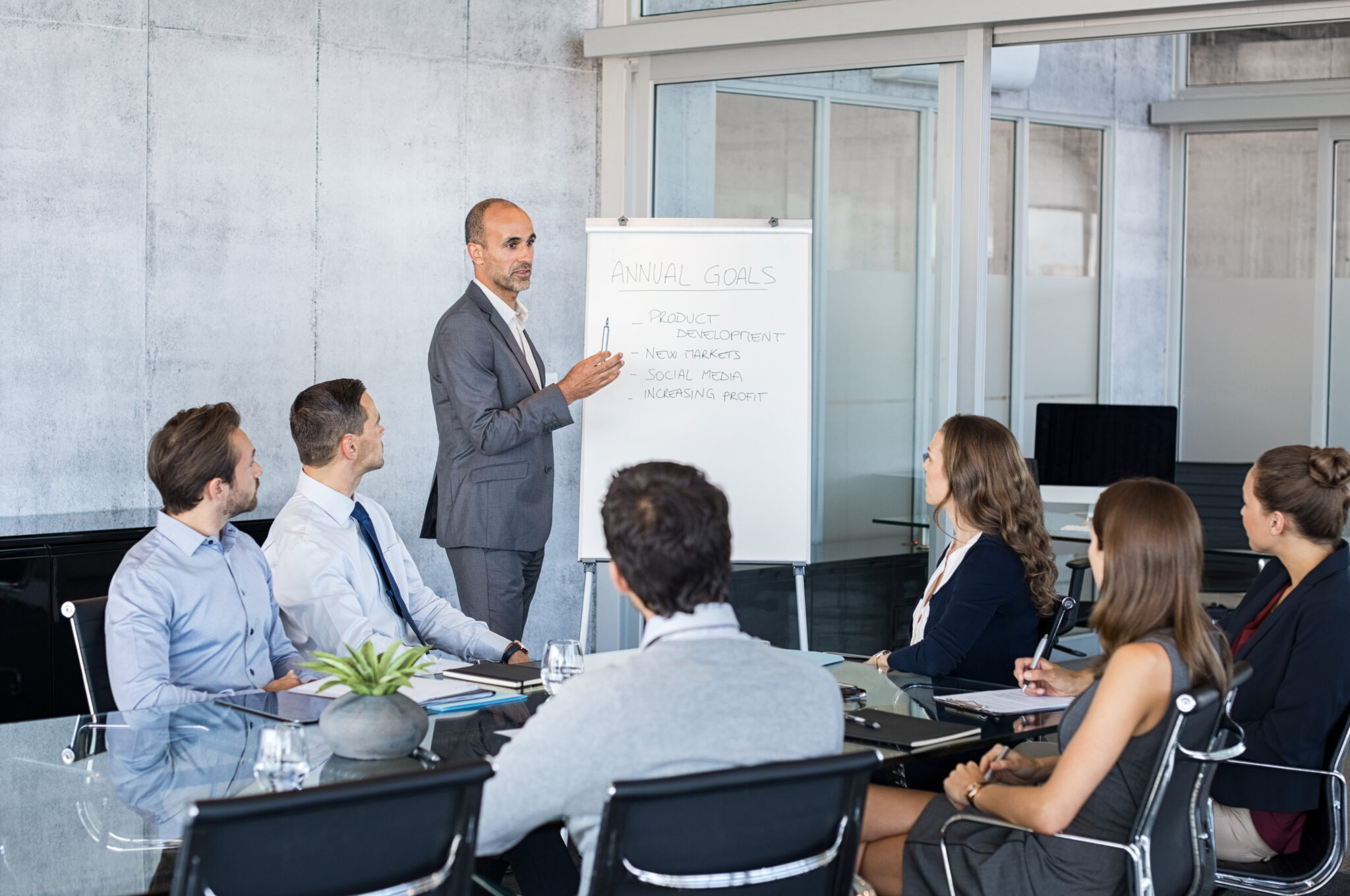 Ein Geschäftsmann im Anzug präsentiert vor einer Gruppe von fünf Personen, die an einem Konferenztisch sitzen. Er steht neben einem Flipchart, auf dem 'Annual Goals' und verschiedene Punkte wie „Product“, „Team Development“, und „New Markets“ notiert sind. Die Zuhörer wirken konzentriert und machen Notizen.