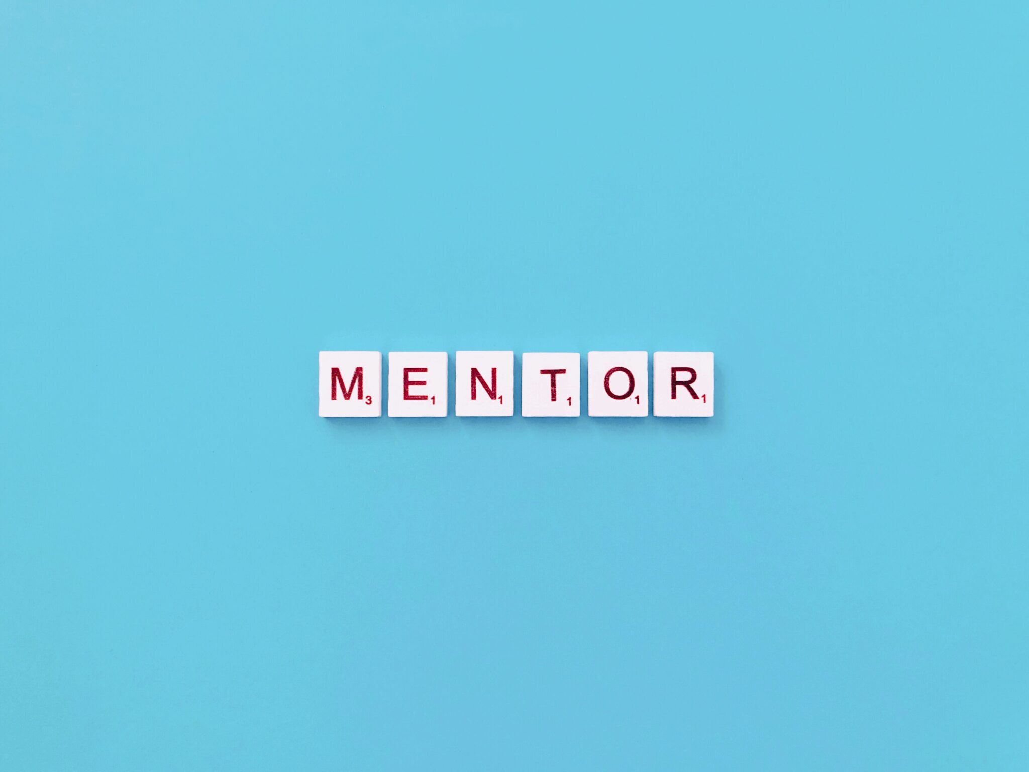 Das Wort „MENTOR“ wird aus weißen Buchstabensteinen vor einem hellblauen Hintergrund gebildet, die an Scrabble-Steine erinnern.