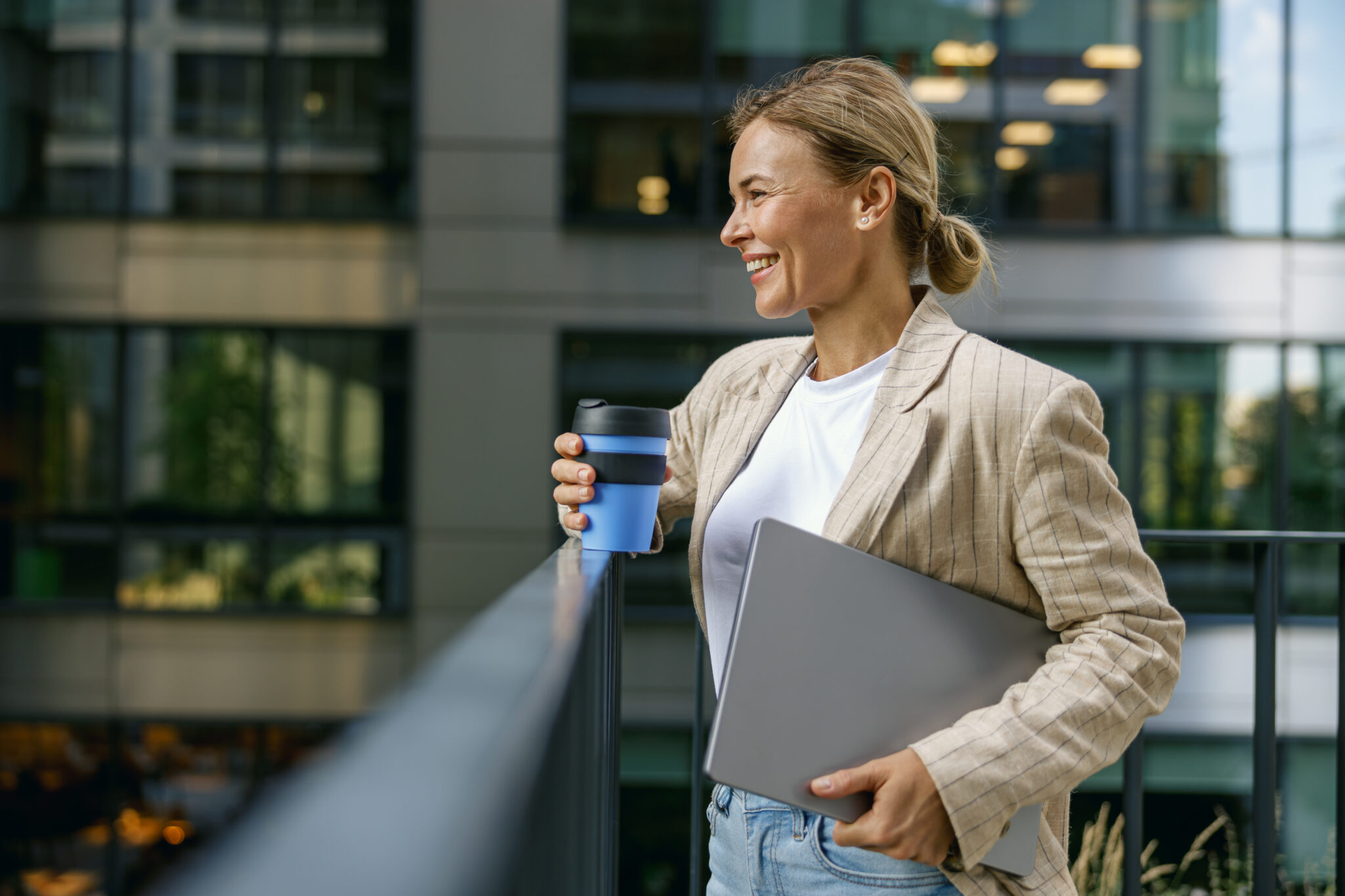 Eine Interim Chief Revenue Officer (CRO) steht auf einem modernen Bürogebäude-Balkon, hält einen Laptop und einen wiederverwendbaren Kaffeebecher in der Hand. Sie trägt einen lässigen Blazer und lächelt, während sie in die Ferne blickt. Das Bild vermittelt Professionalität und Flexibilität, passend für eine CRO-Position, die interimistisch besetzt wird, bis eine dauerhafte Lösung gefunden oder intern aufgebaut ist.