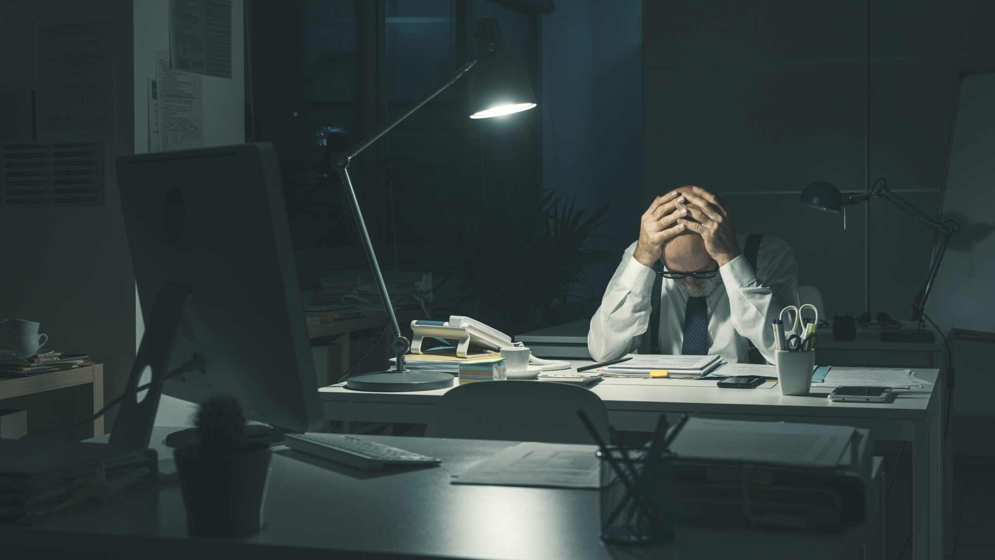 Eine männliche Führungskraft sitzt in einem dunklen Büro und hält sich erschöpft den Kopf mit beiden Händen. Auf dem Schreibtisch liegen Dokumente und ein Computerbildschirm ist zu sehen.