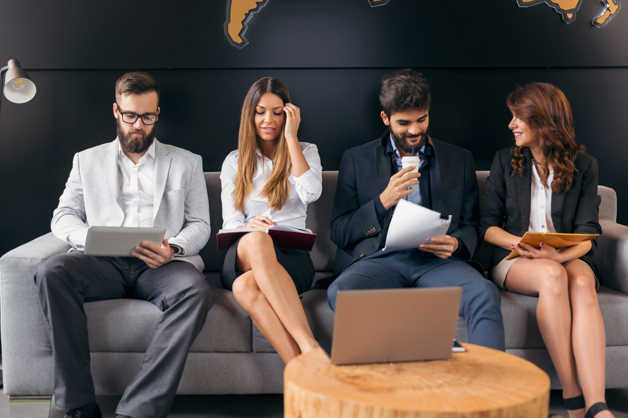 Vier professionelle Personen in Businesskleidung sitzen auf einem Sofa und warten auf ihr Bewerbungsgespräch. Sie beschäftigen sich mit Laptops, Dokumenten und Notizen.