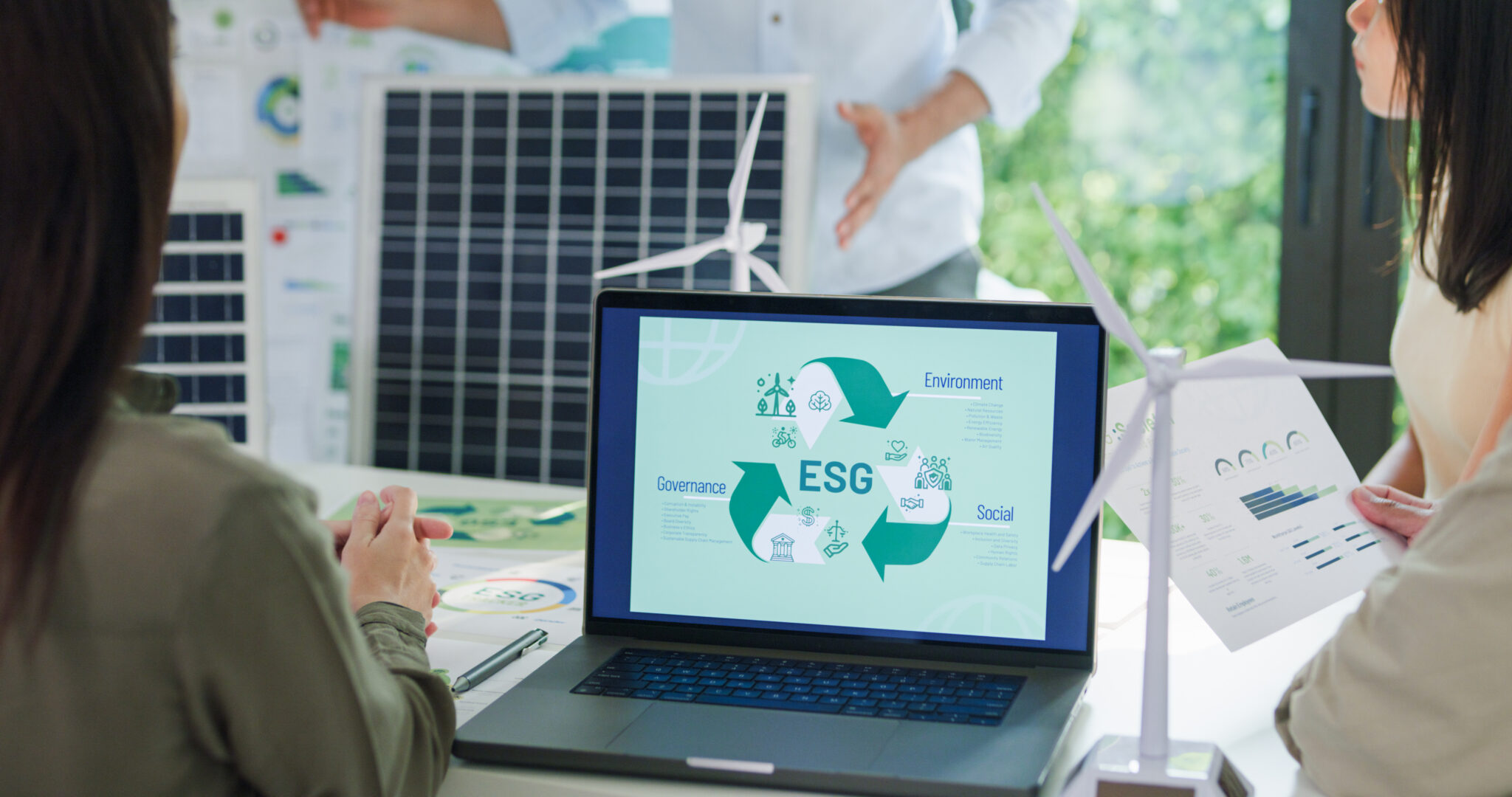 In einem Besprechungsraum arbeiten zwei Personen an einem Laptop, der ein ESG-Diagramm zeigt. ESG steht für Environment (Umwelt), Social (Soziales) und Governance (Unternehmensführung). Im Hintergrund sind Solarpanele und ein Modell einer Windturbine zu sehen, die auf nachhaltige Energie hinweisen. Eine Person hält ein Dokument mit Datenvisualisierungen.