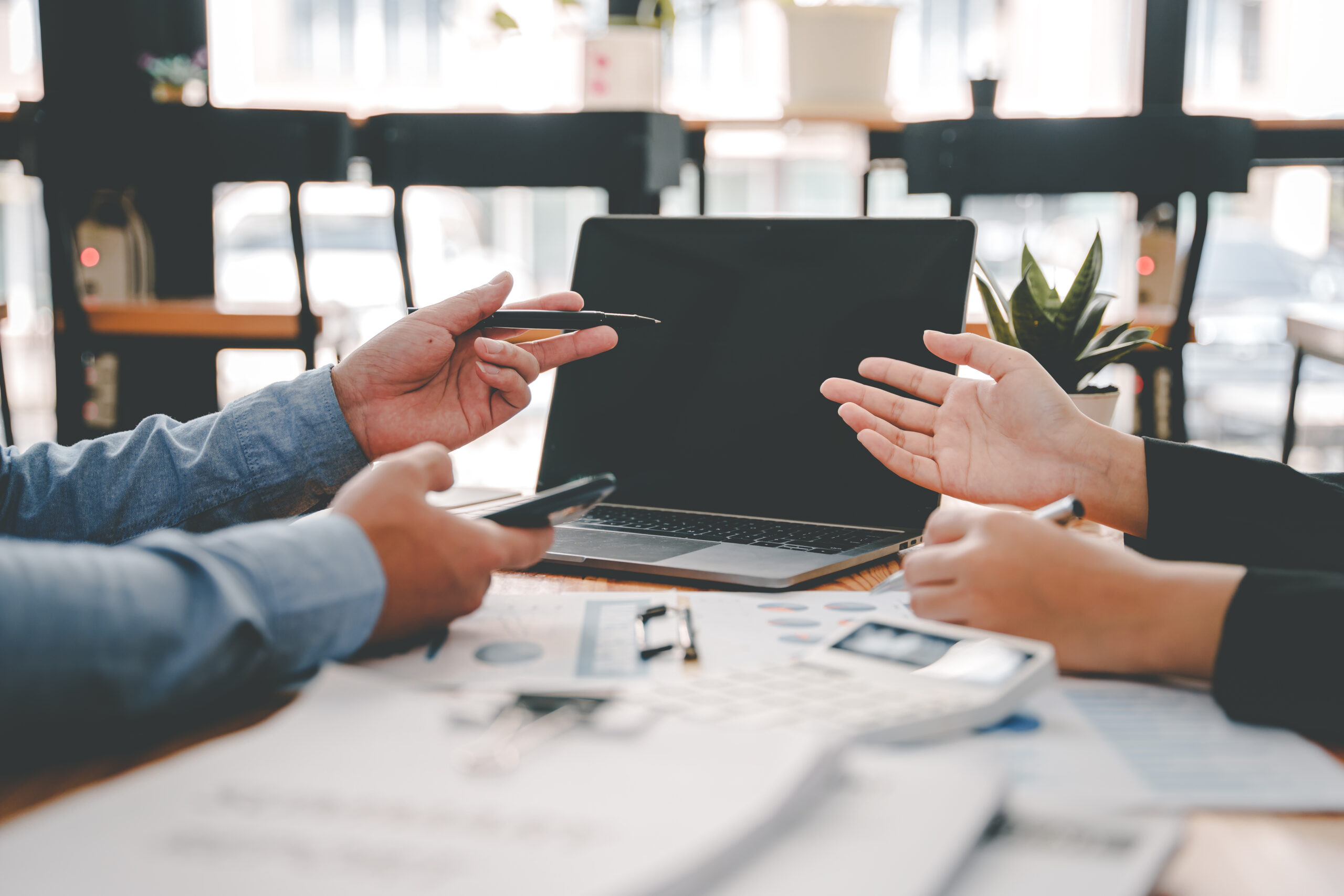 Zwei Personen in einem Kundengespräch in einer Bankfiliale, die über Finanzunterlagen diskutieren. Eine Person hält ein Smartphone, während die andere mit einer erklärenden Handgeste antwortet. Auf dem Tisch liegen Dokumente, ein Taschenrechner und ein Laptop, was auf eine Beratungssituation zu Finanz- oder Kreditangelegenheiten im Bankwesen hindeutet.