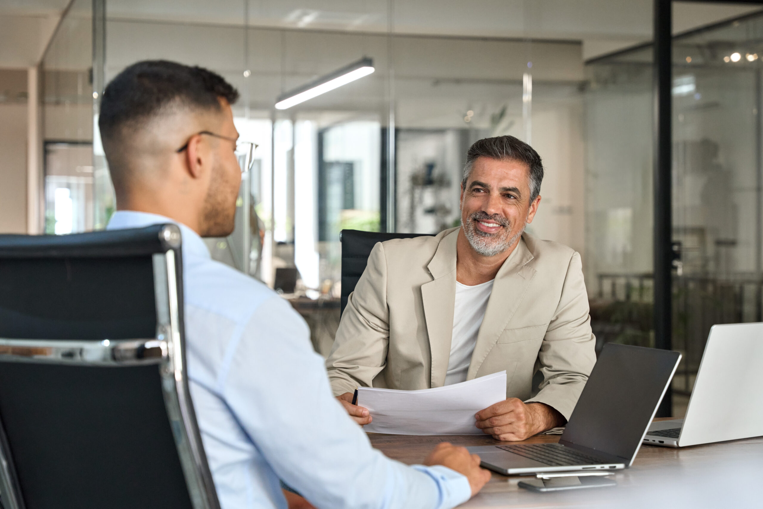 Zwei Männer in einem formellen Bewerbungsgespräch in einem modernen Büro. Einer der Männer, im beigen Anzug, lächelt und hält ein Blatt Papier, während der andere ihm gegenübersitzt und aufmerksam zuhört. Auf dem Tisch stehen zwei Laptops. Das Bild vermittelt eine professionelle Interviewsituation zwischen einem Personalberater und einem Kandidaten für die Position eines Vorstands Marktfolge.