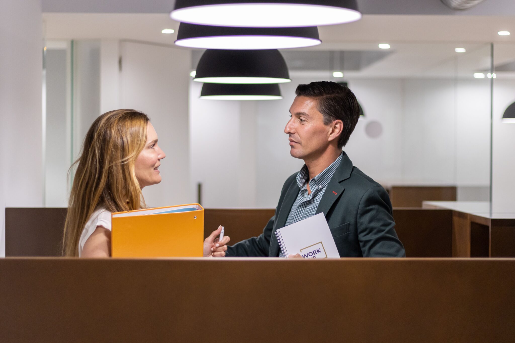 Zwei Geschäftspersonen unterhalten sich in einem modernen Büro, halten Unterlagen und Notizbücher in den Händen, was die Wichtigkeit der Zusammenarbeit der Omnikanalsteuerung mit anderen Abteilungen hervorhebt.