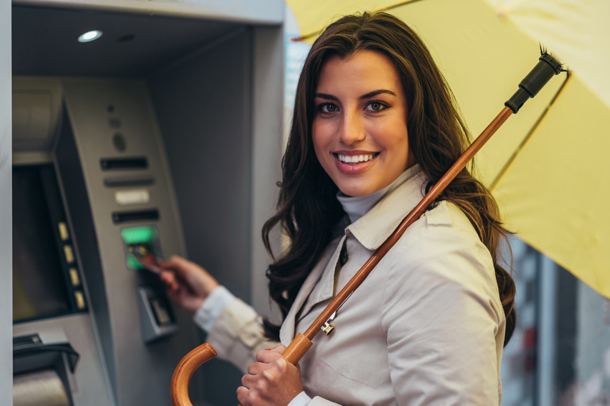 Eine lächelnde Frau steht mit einem gelben Regenschirm vor einem Geldautomaten und nutzt ihre Bankkarte, was den Fokus auf Kundenbedürfnisse als Grundlage jeder Omnikanalstrategie verdeutlicht.