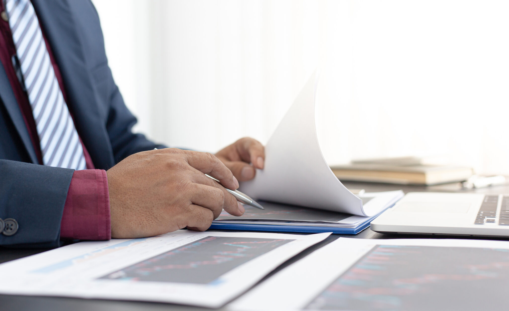 Symbolbild für den Karriereweg des COO: Eine Person in Business-Kleidung blättert durch Dokumente auf einem Schreibtisch, umgeben von Unterlagen, Diagrammen und einem Laptop. Die Szene betont die Bedeutung von Ausbildung und Studium sowie analytischen Fähigkeiten und strategischem Denken für eine Karriere als Chief Operating Officer.