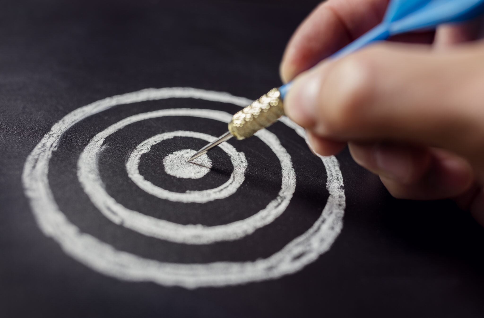 Eine Hand zielt mit einem Dartpfeil auf die Mitte einer Kreide-Zielscheibe auf einer schwarzen Tafel, symbolisierend für präzise Strategien im Executive Search in Frankfurt.