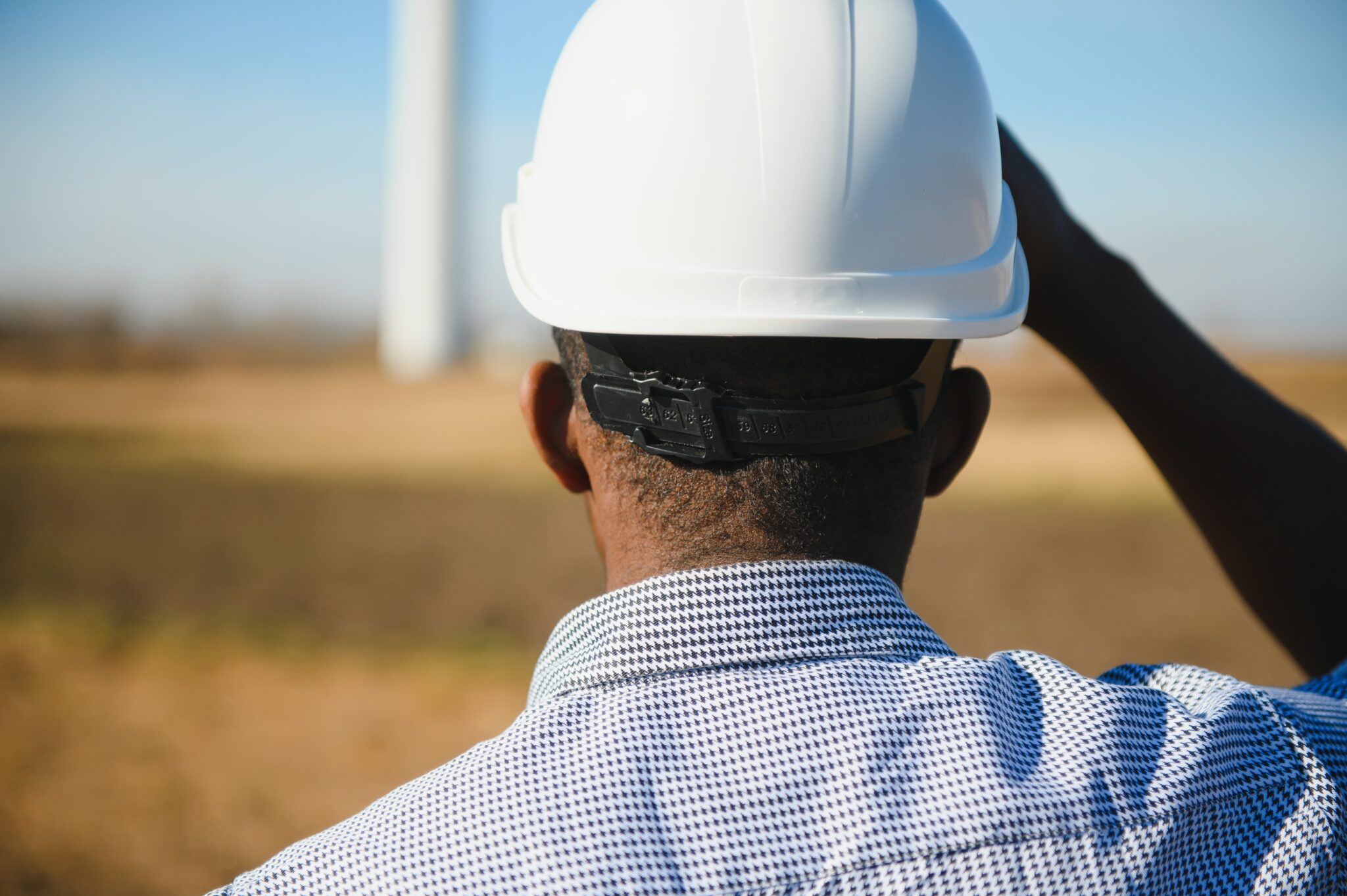 Ein Mann mit Schutzhelm und kariertem Hemd betrachtet eine offene Landschaft, im Hintergrund ist unscharf eine Windkraftanlage zu erkennen. Der Untertitel „Headhunter für Energiewirtschaft finden Ihre gesuchten Fachkräfte“ hebt die Unterstützung durch spezialisierte Personalberater bei der Rekrutierung von Fachkräften für die Energiebranche hervor. Die Szene symbolisiert die Verbindung von Fachkenntnissen und nachhaltiger Energieerzeugung.