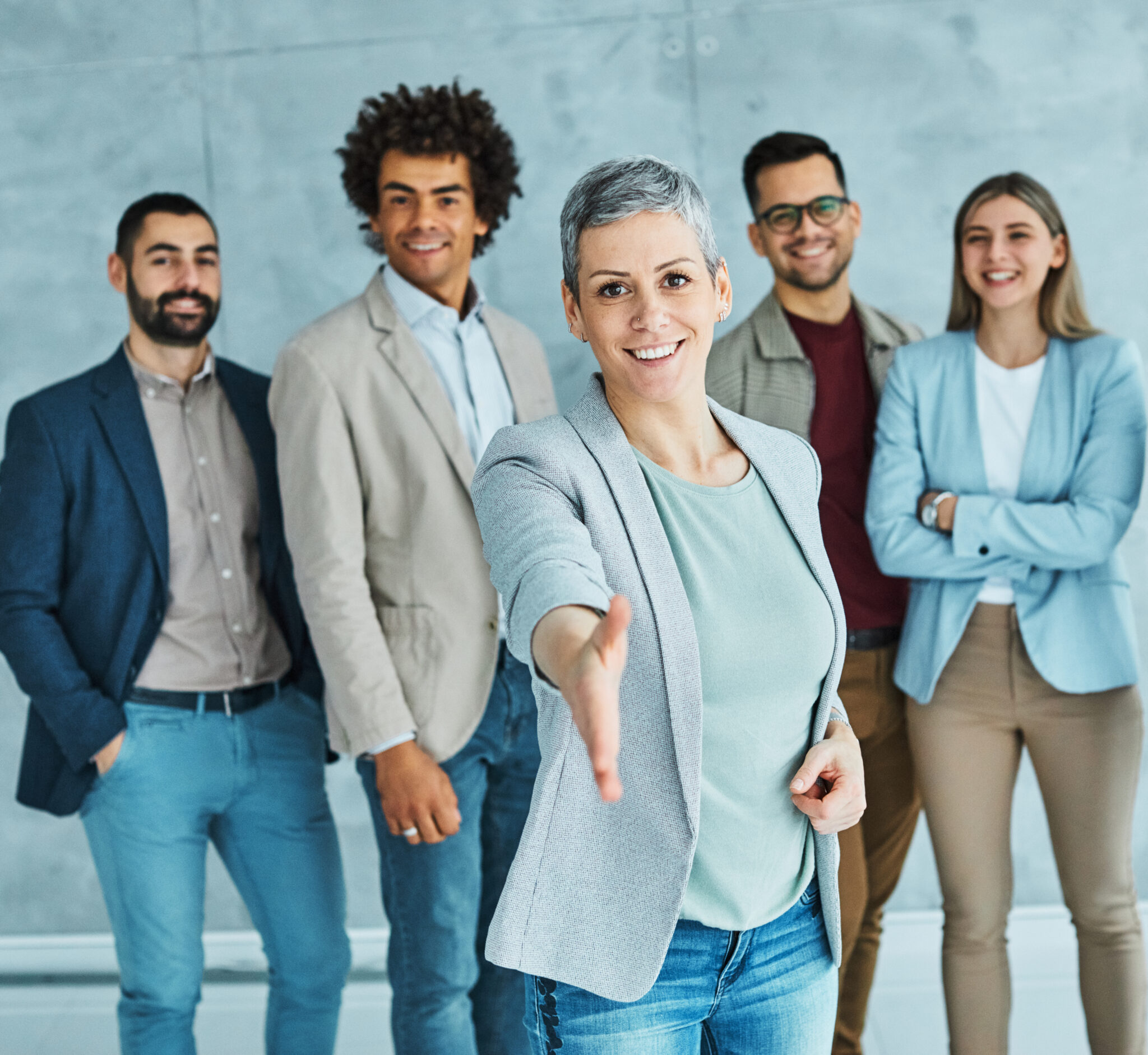 Gruppe von lächelnden, diversen Mitarbeitenden, eine Frau streckt die Hand aus zur Begrüßung. Darstellung eines attraktiven Arbeitgebers durch flexible Arbeitsmodelle.