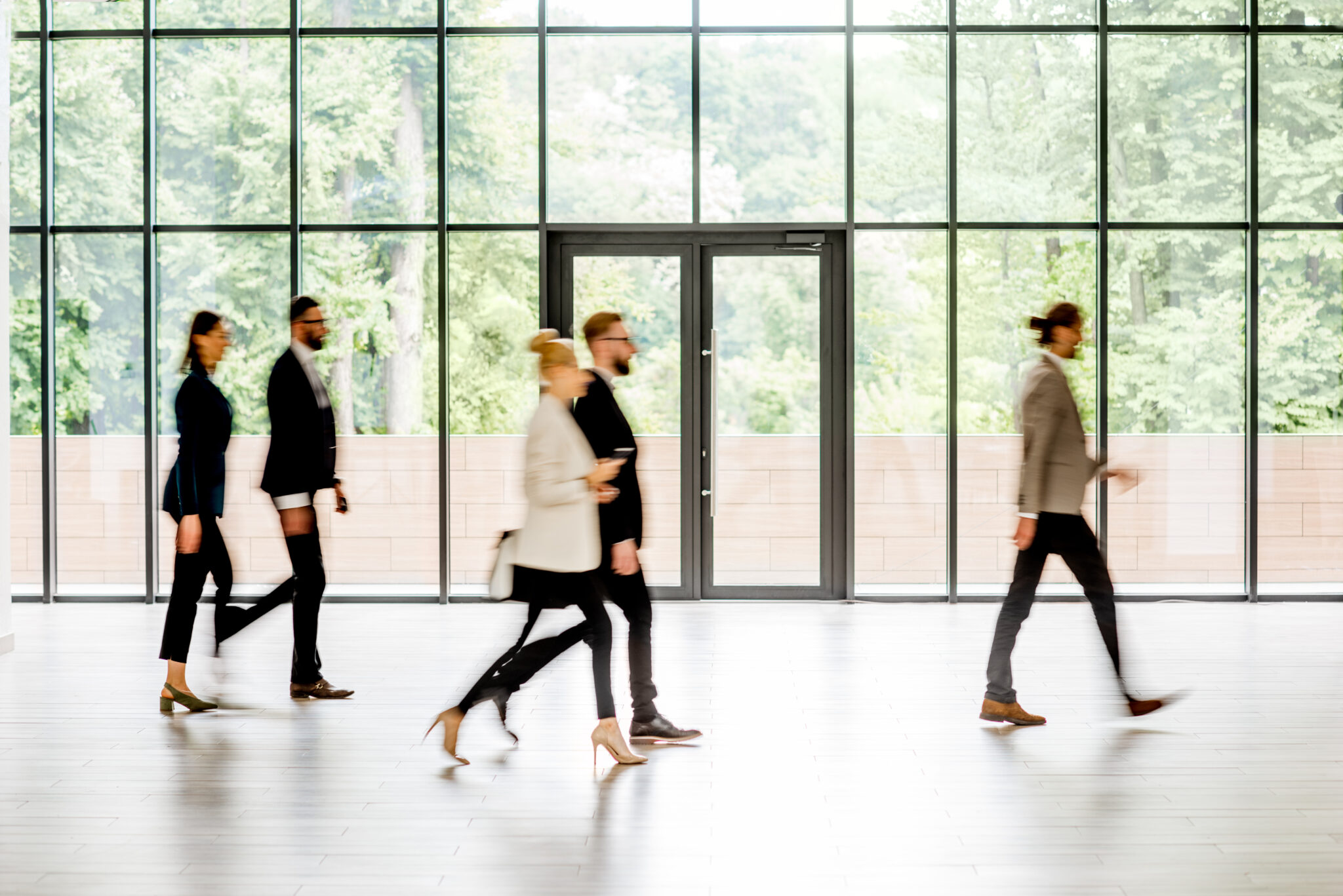 Fotografie in einem modernen, lichtdurchfluteten Raum mit großen Glasfenstern. Mehrere Personen in Businesskleidung gehen dynamisch durch den Raum. Die Szenerie vermittelt Bewegung und Geschäftigkeit. Im Hintergrund ist eine grüne Landschaft sichtbar.