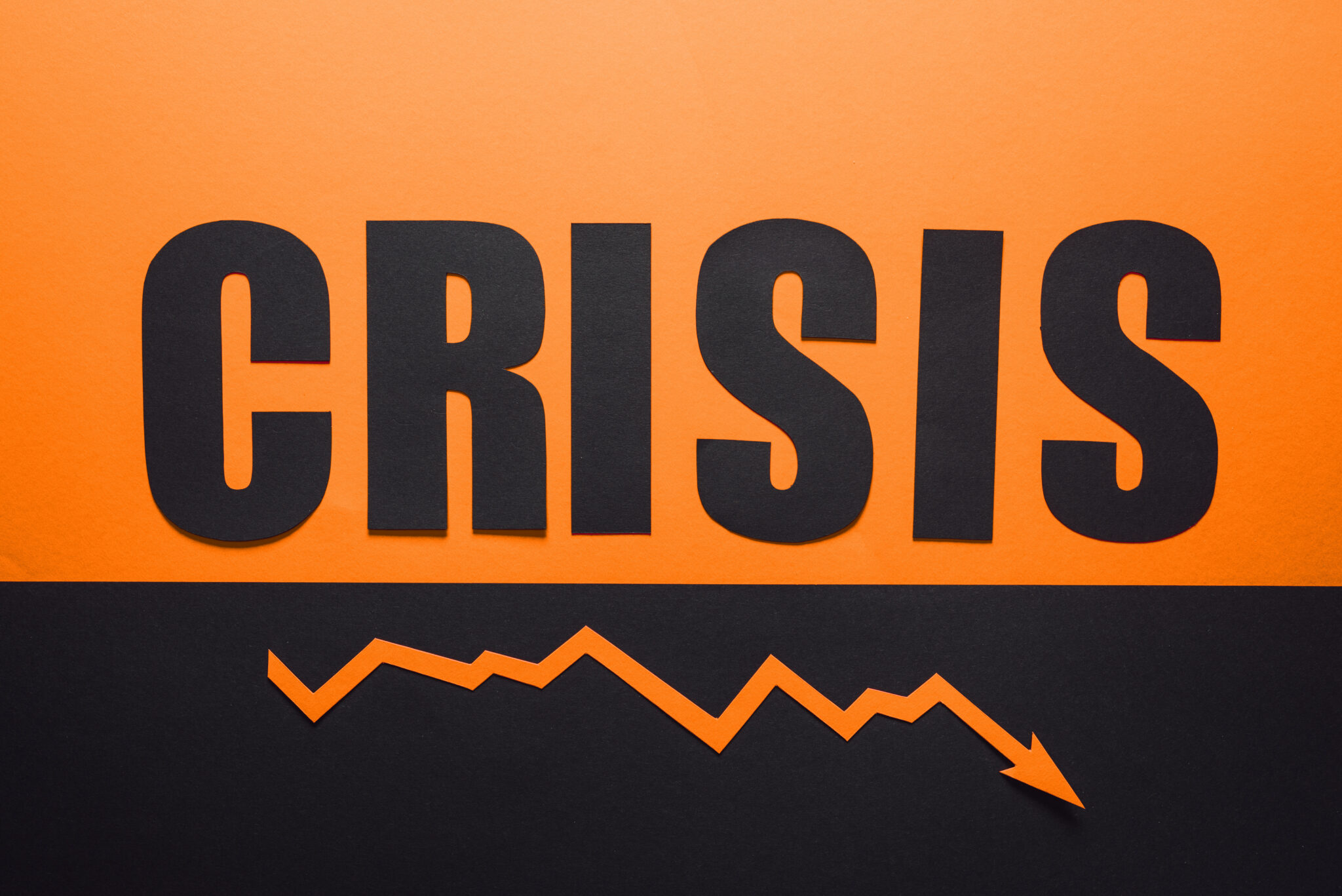 Bild mit dem Wort 'CRISIS' in großen schwarzen Buchstaben auf einem zweifarbigen Hintergrund, oben orange und unten schwarz. Unter dem Wort verläuft ein Zickzack-Pfeil, der einen fallenden Trend darstellt.