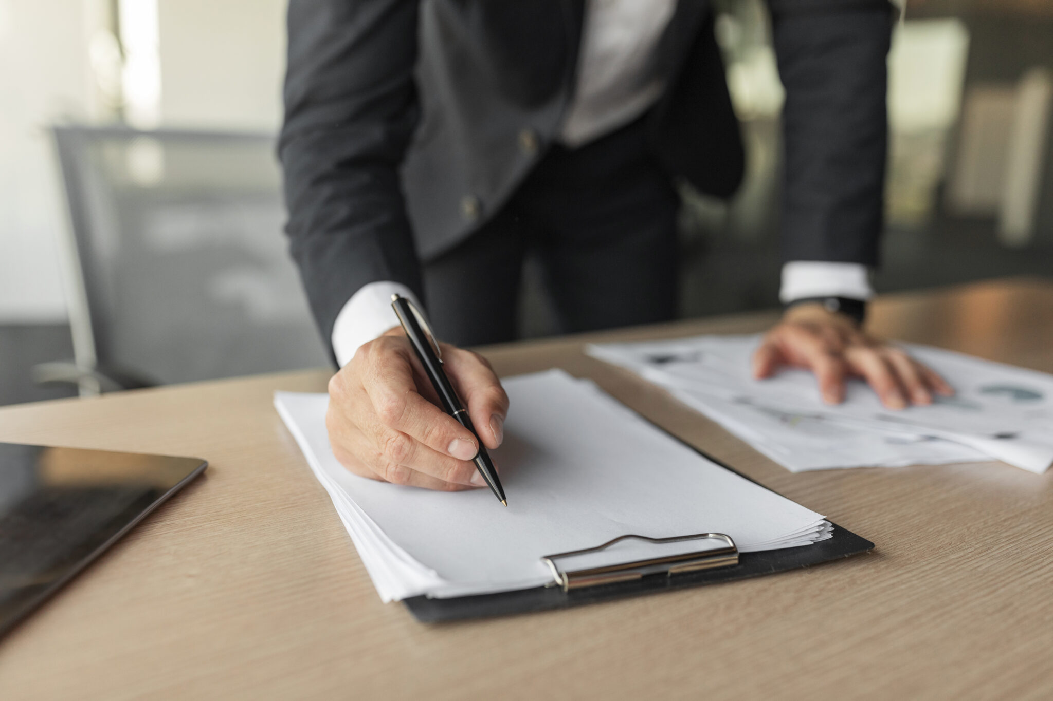 Fotografie eines Interim Managers in einem dunklen Anzug, die sich über einen Tisch beugt und mit einem Stift einen Vertrag auf einem Klemmbrett unterschreibt. Im Hintergrund liegen weitere Unterlagen auf dem Tisch.