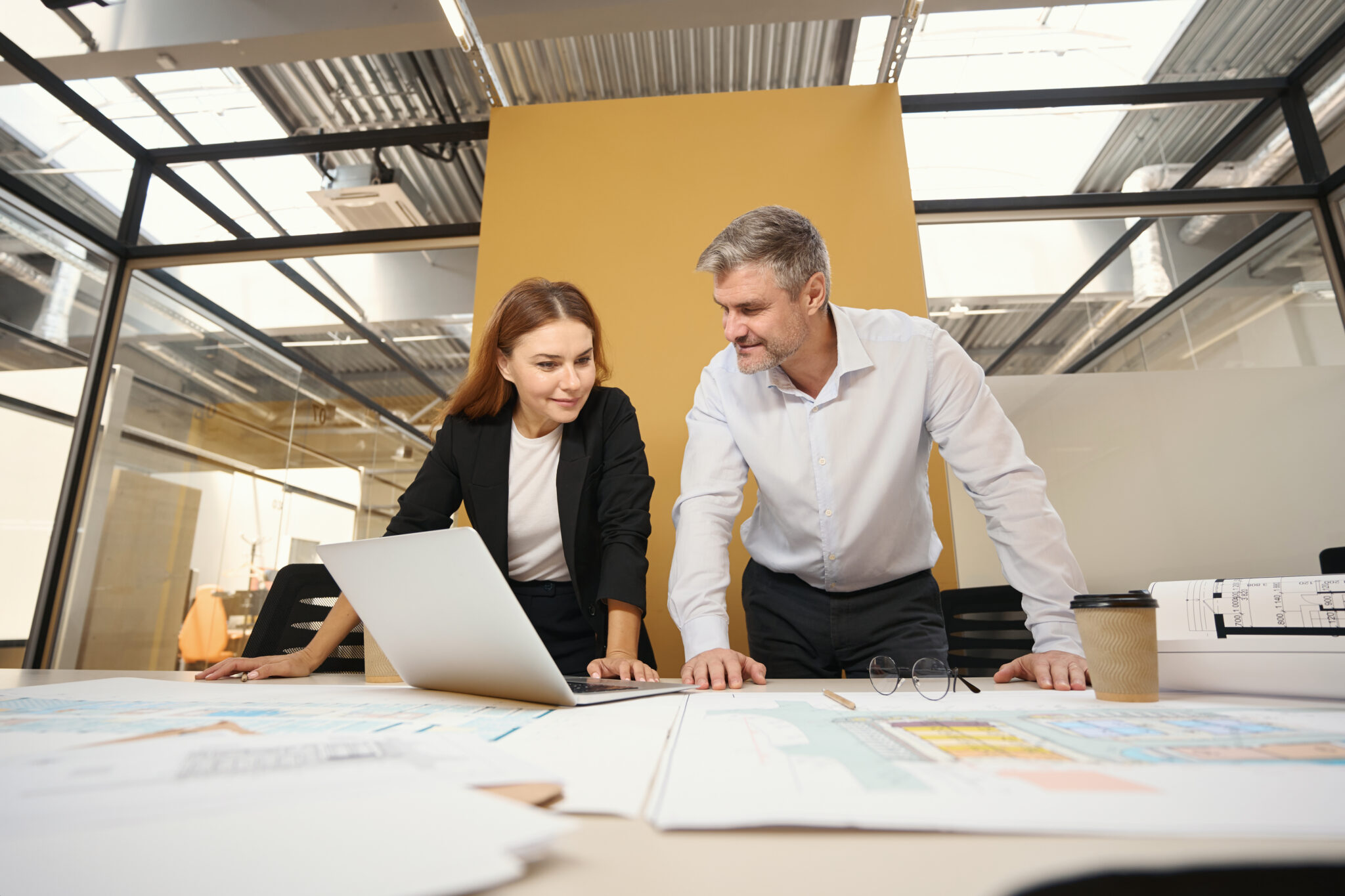 Fotografie einer Mitarbeiterin und eines Interim Managers in einem modernen Büro mit Glaswänden und Industriedecke. Beide stehen an einem Tisch mit Plänen und Dokumenten, während sie gemeinsam auf einen Laptop schauen. Die Mitarbeiterin trägt ein schwarzes Blazer-Outfit, der Interim Manager ein weißes Hemd. Auf dem Tisch stehen auch eine Brille und ein Kaffeebecher.