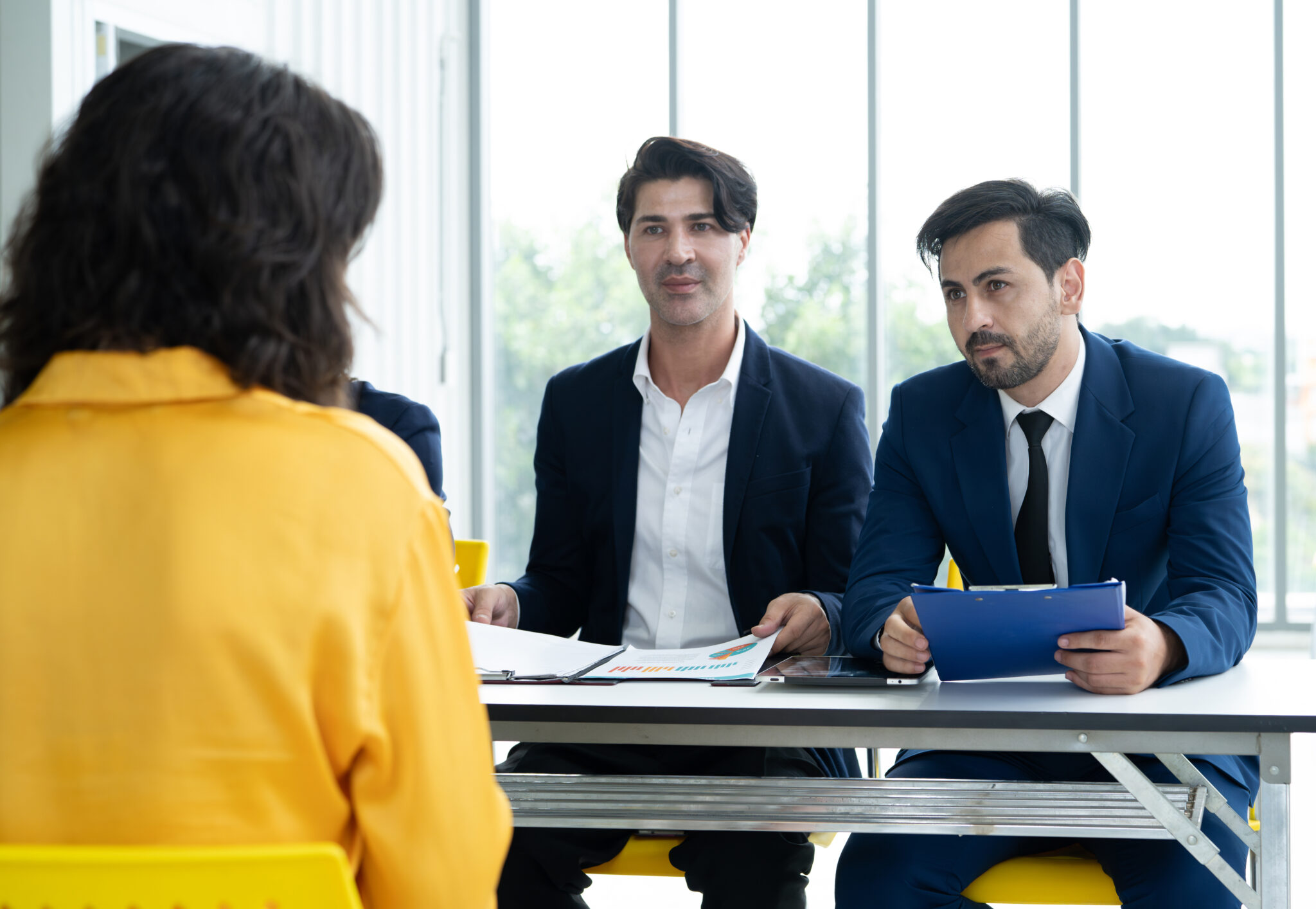 Drei Headhunter in Business-Anzügen sitzen an einem Tisch und führen ein Gespräch mit einer Bewerberin in gelber Kleidung, was den Einsatz von Headhuntern im Fachkräftemangel symbolisiert.