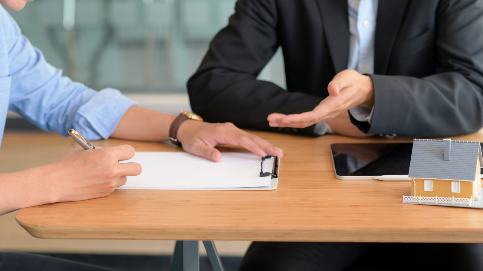 Ein Kreditanalyst und ein Kunde sitzen an einem Tisch. Der Kunde füllt ein Formular auf einem Klemmbrett aus, während ein kleines Modellhaus und ein Tablet auf dem Tisch liegen.