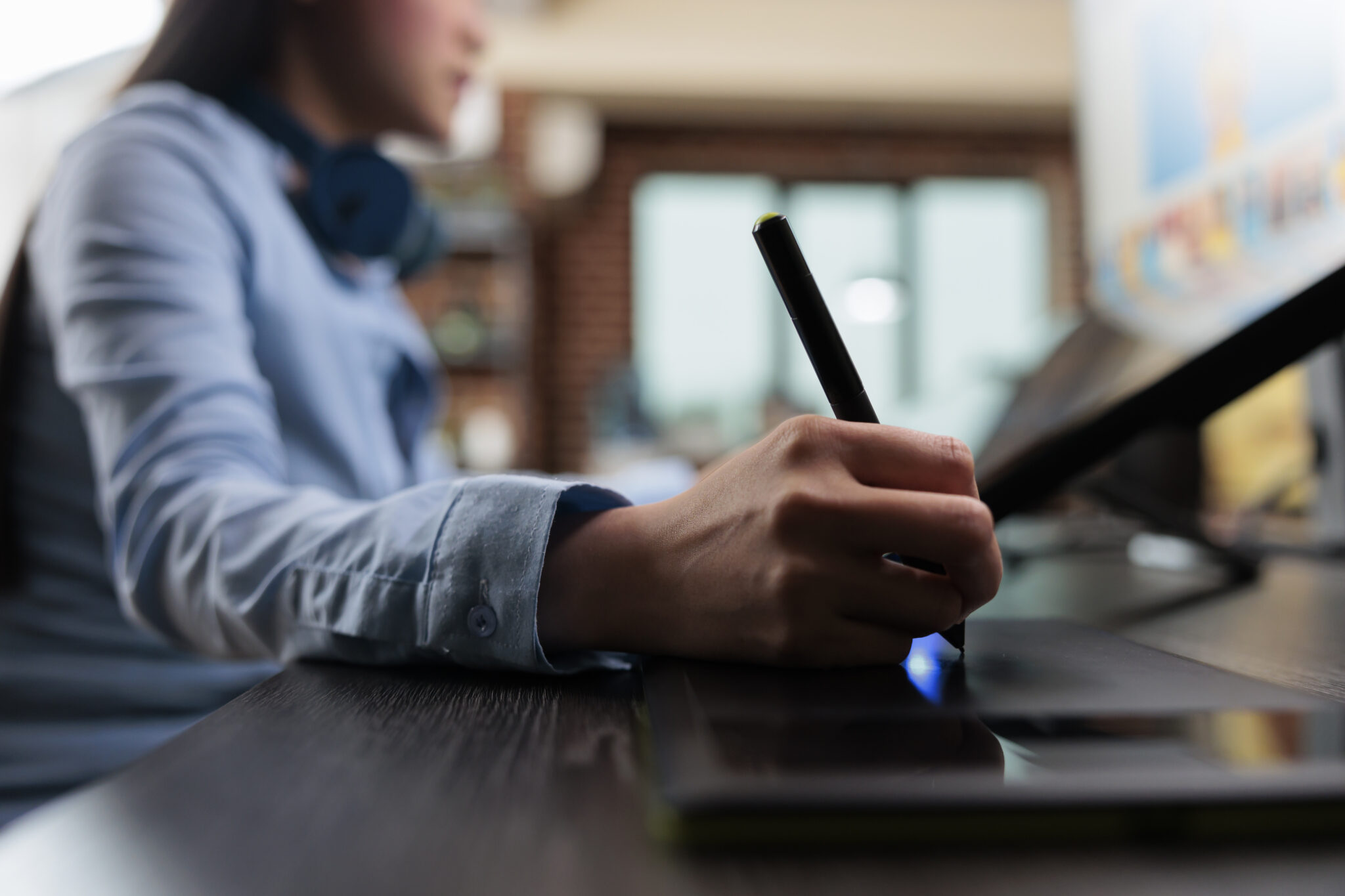 Eine Mitarbeiterin eines Maschinenbauers im Homeoffice schreibt mit einem digitalen Stift auf ein Grafiktablett, während ein Laptop vor ihr steht. Sie trägt ein Hemd und hat Kopfhörer um den Hals. Im Hintergrund ist ein Fenster mit unscharfem Ausblick zu sehen.