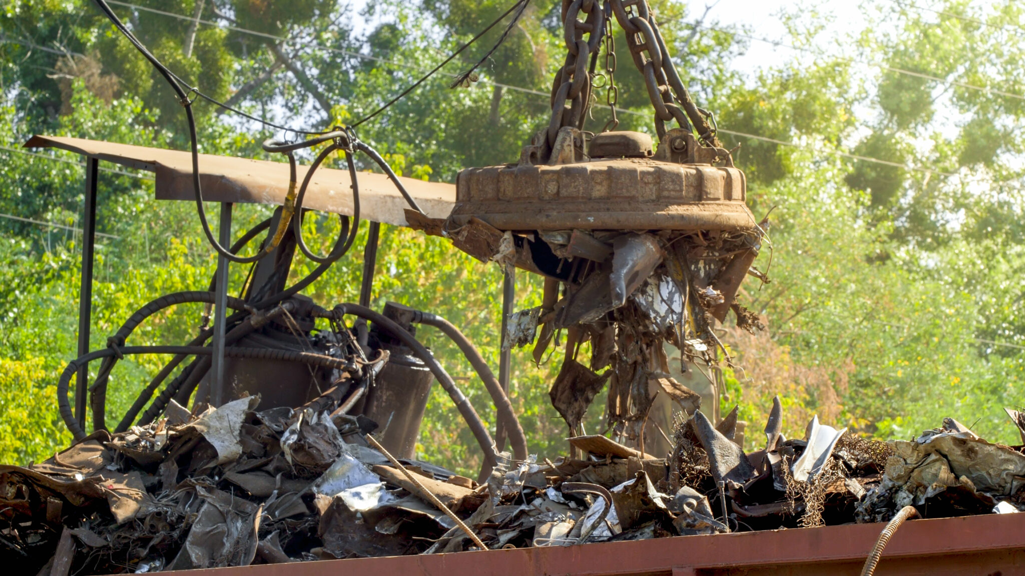 Magnetkran hebt Metallteile auf einem Schrottplatz. Im Hintergrund grüne Bäume. Symbolbild für Metallrecycling, Rohstoffwiederverwertung und nachhaltige Industrieprozesse.