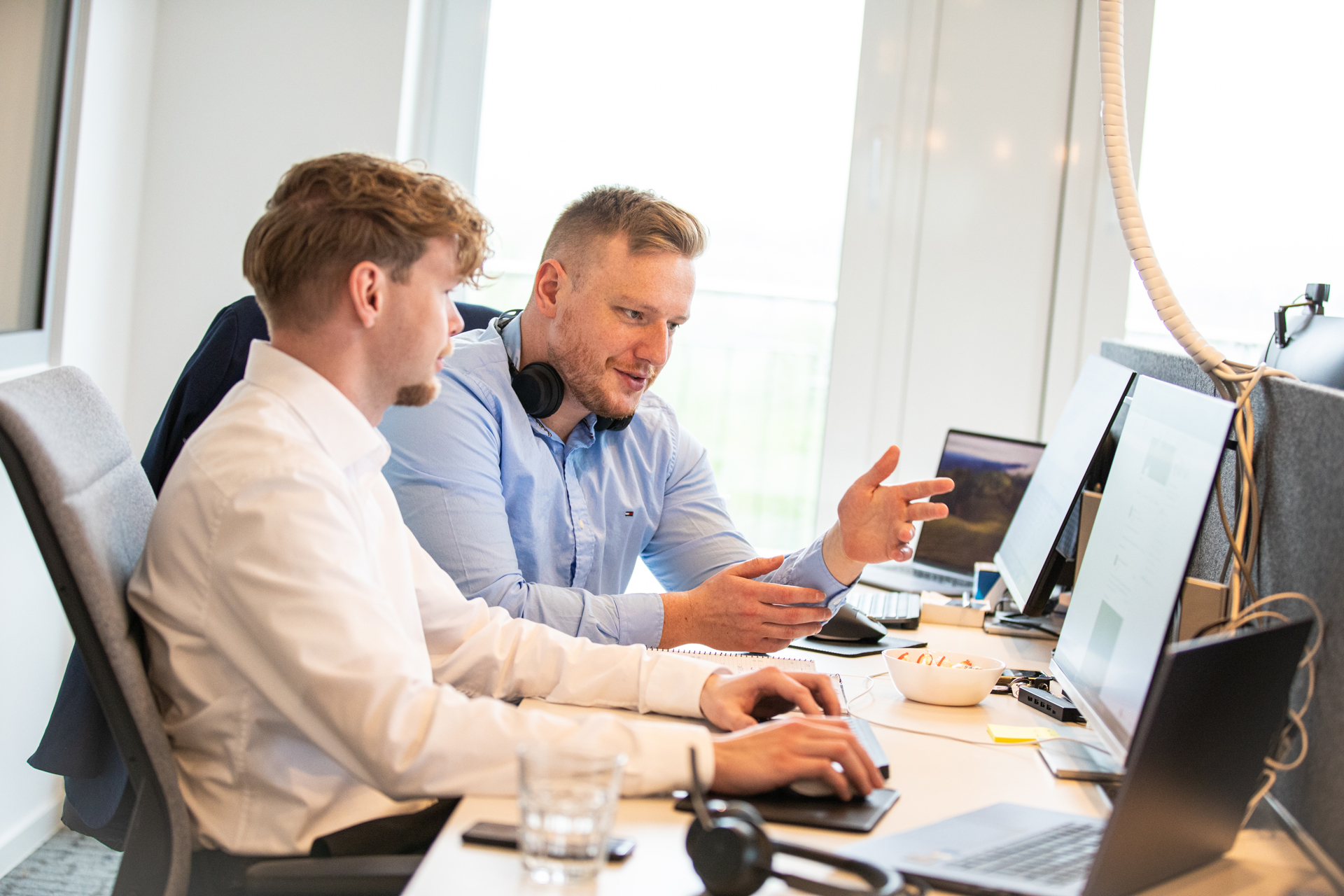 Zwei männliche Personalberater in Business-Kleidung arbeiten gemeinsam an einem Computer in einem modernen Büro. Einer erklärt gestikulierend, während der andere konzentriert zuhört.