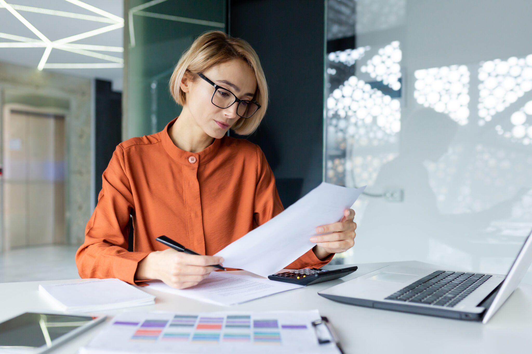 Junge Frau mit Brille und kurzem blondem Haar sitzt an einem modernen Schreibtisch in einem gut beleuchteten Büro. Sie trägt eine orangefarbene Bluse und überprüft konzentriert ein Dokument, während sie mit einem Stift in der Hand Notizen macht. Auf dem Schreibtisch liegen weitere Unterlagen, ein Laptop, ein Taschenrechner und ein Tablet. Der Hintergrund zeigt ein stilvolles Bürodesign mit Glaswänden und geometrischen Lichtelementen.