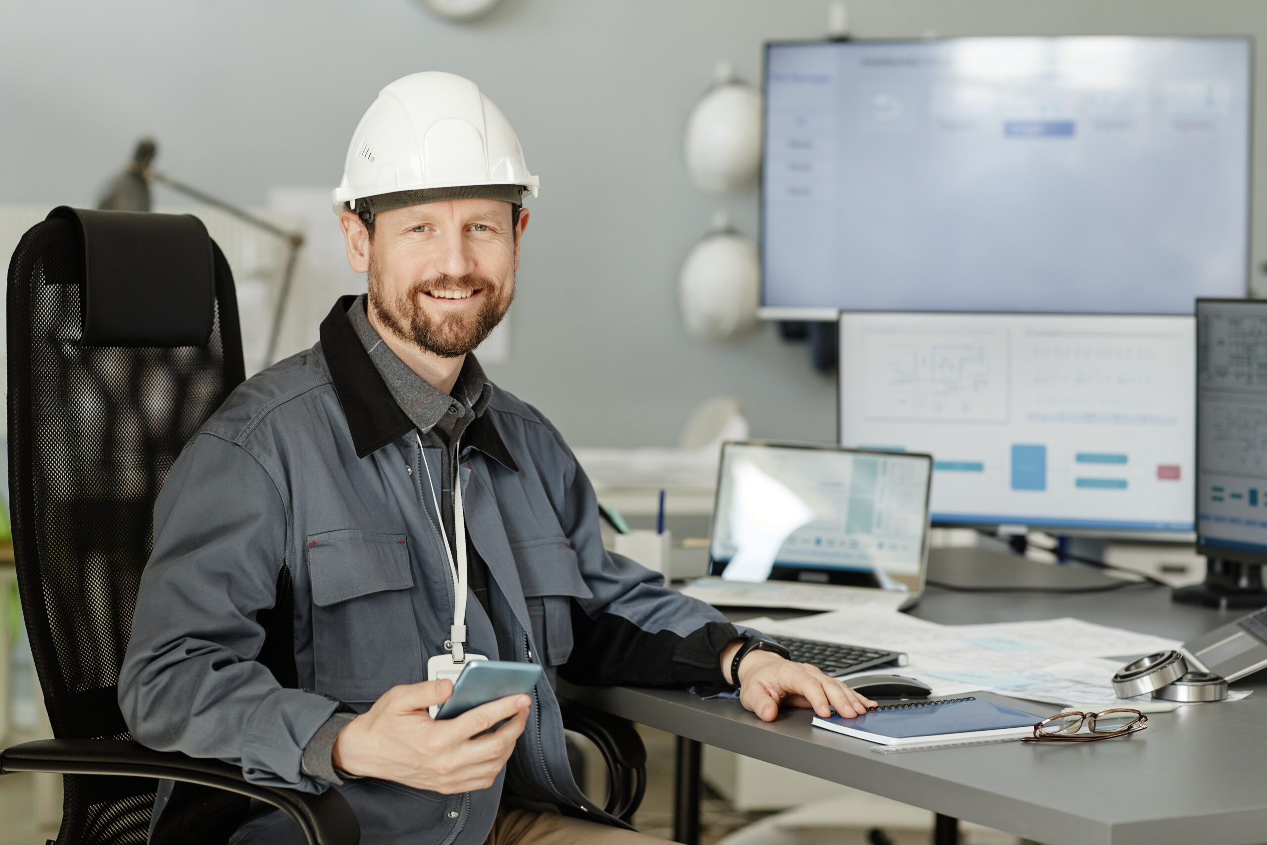 Freundlicher Projektleiter mit Helm und Smartphone am Schreibtisch im Büro.