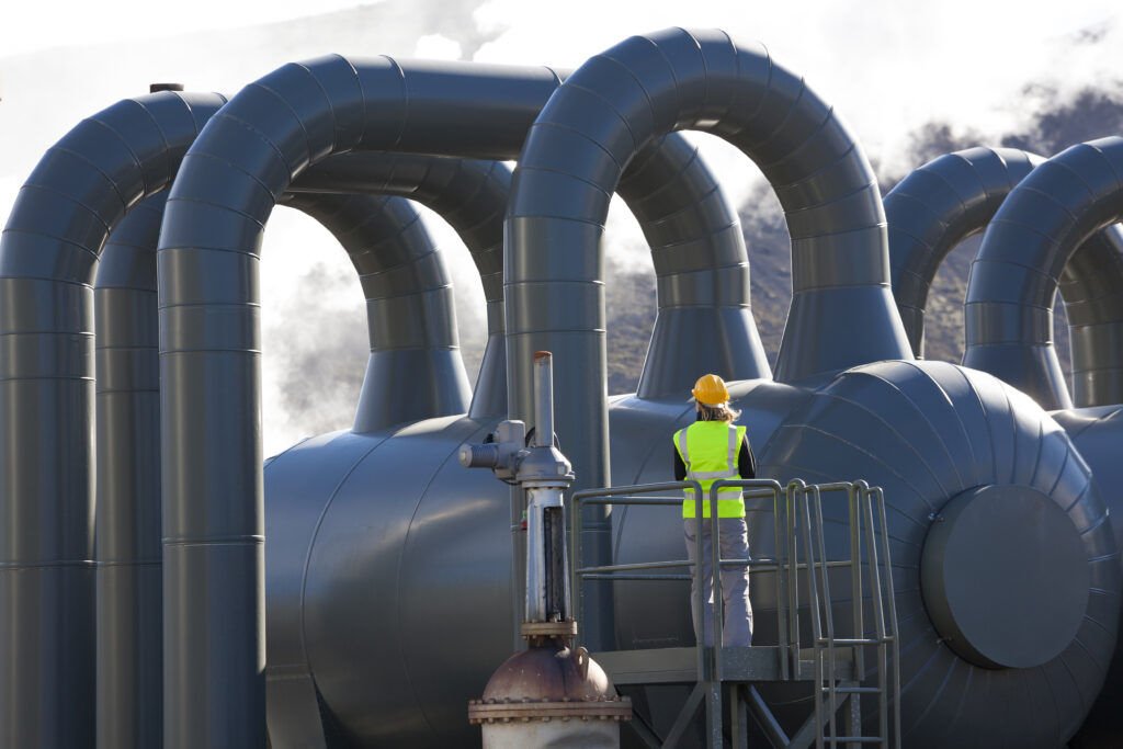 Eine Person mit Schutzhelm und Warnweste steht auf einer Plattform und blickt auf große, gewölbte Rohre einer industriellen Geothermie-Anlage, aus denen Dampf aufsteigt.