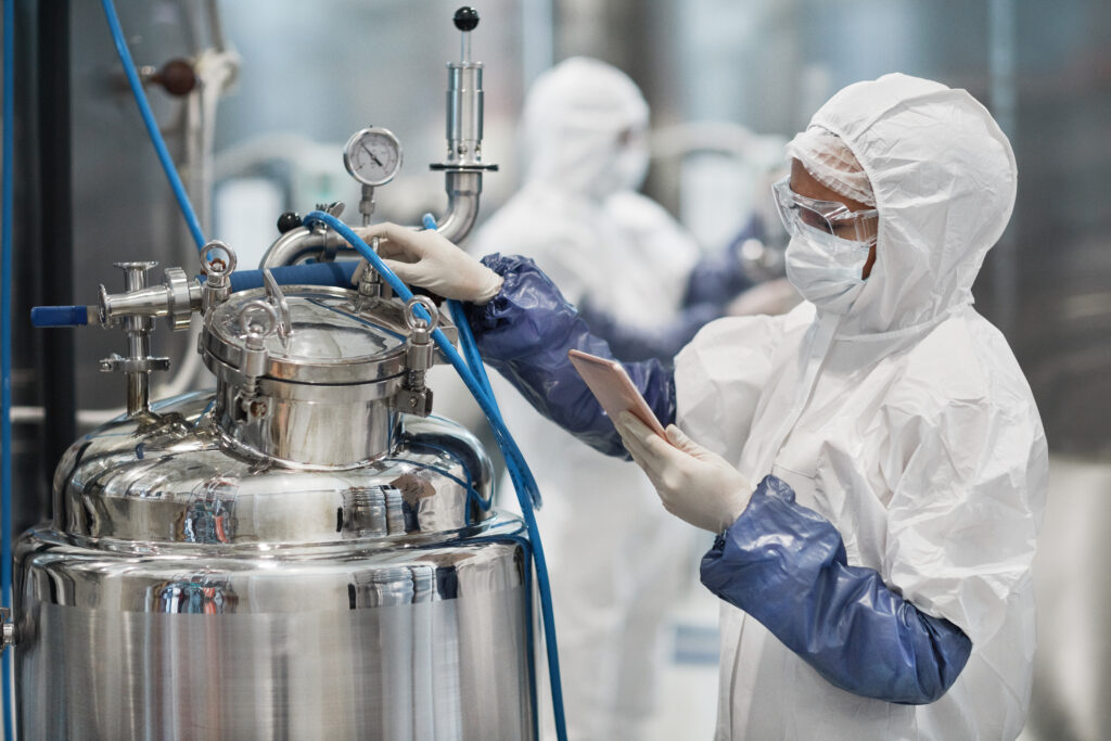 Zwei Personen in weißen Schutzanzügen mit Kapuzen, Gesichtsmasken und blauen Gummihandschuhen arbeiten an einem großen, glänzenden Kessel in einer industriellen Anlage. Die Person im Vordergrund bedient ein Tablet.