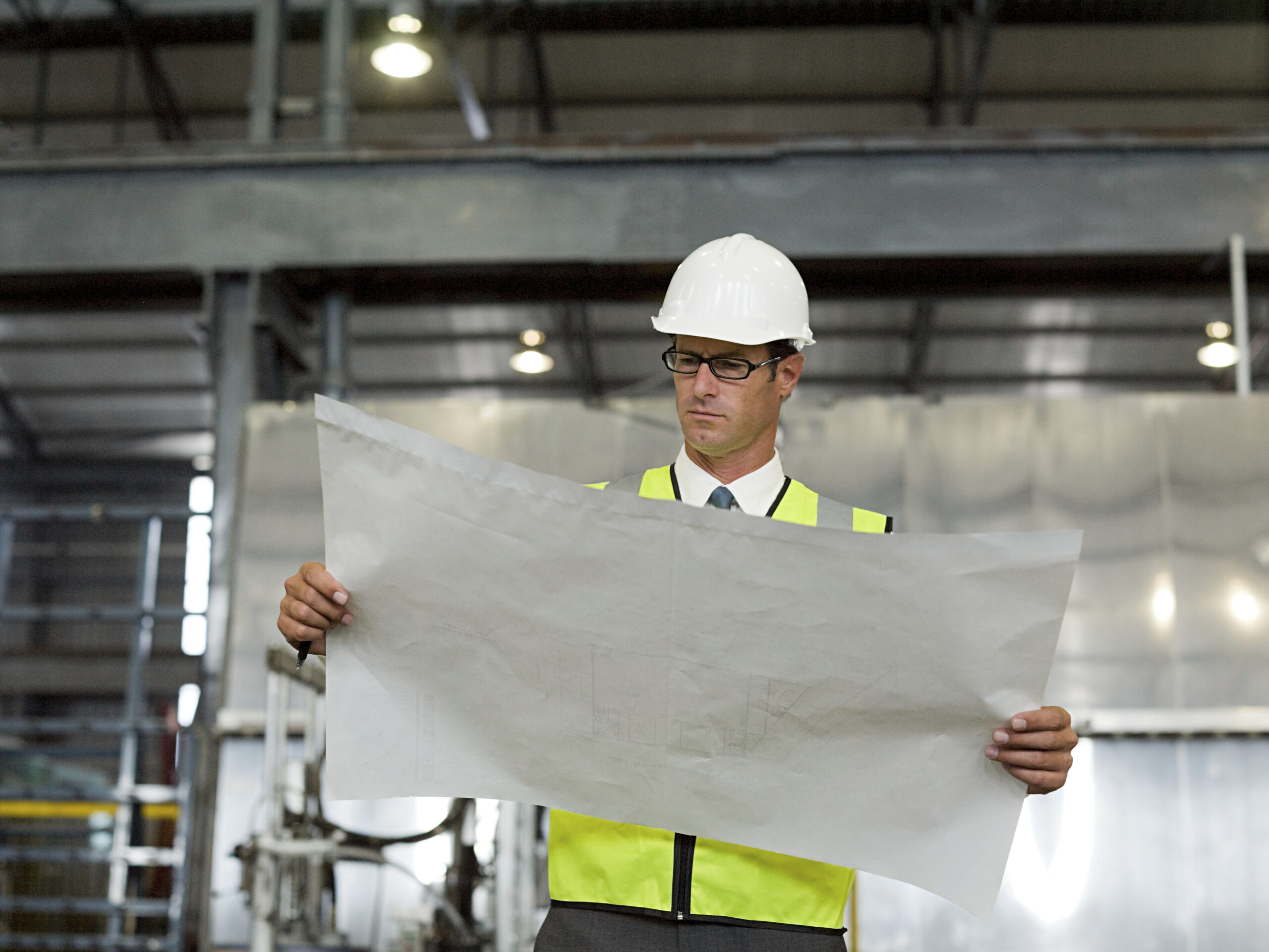 REFA-Ingenieur in Schutzweste und Helm studiert Bauplan in einer Fabrikhalle.